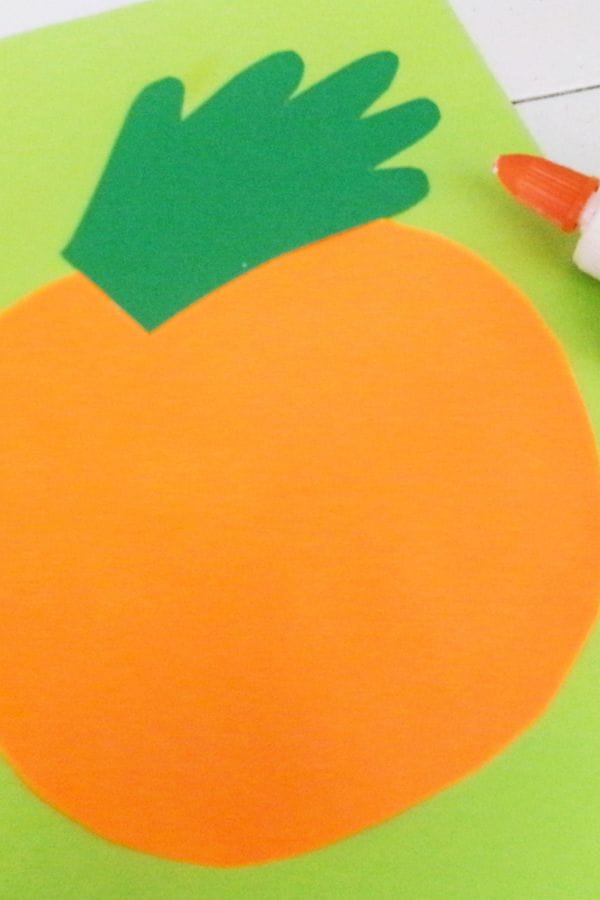 An orange circle with a green hand-shaped cutout and glue stick on green paper, resembling a simple Handprint Pumpkin Craft.