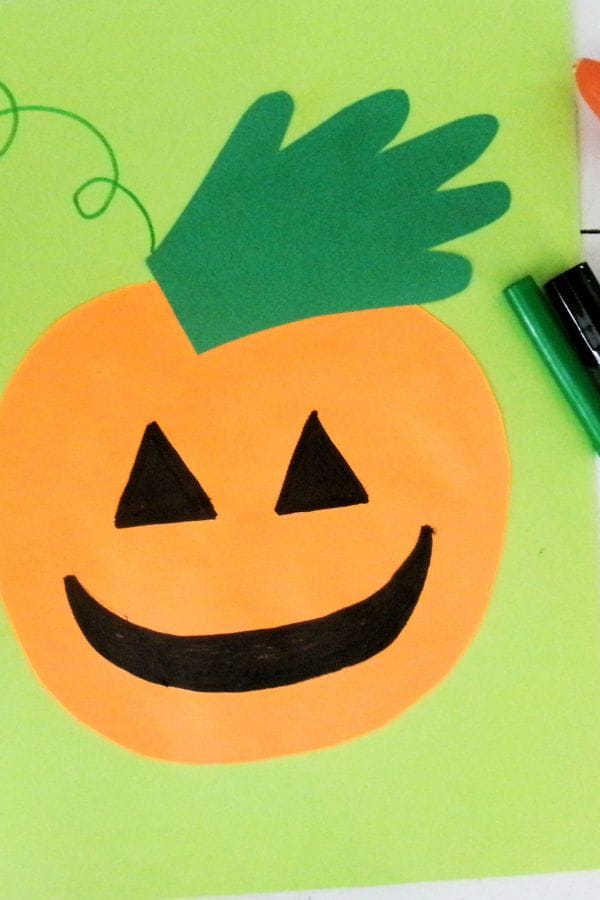 A handprint pumpkin craft featuring a paper cutout pumpkin with a green stem, black triangle eyes, and a smiling mouth, arranged on a light green background with a green marker nearby.