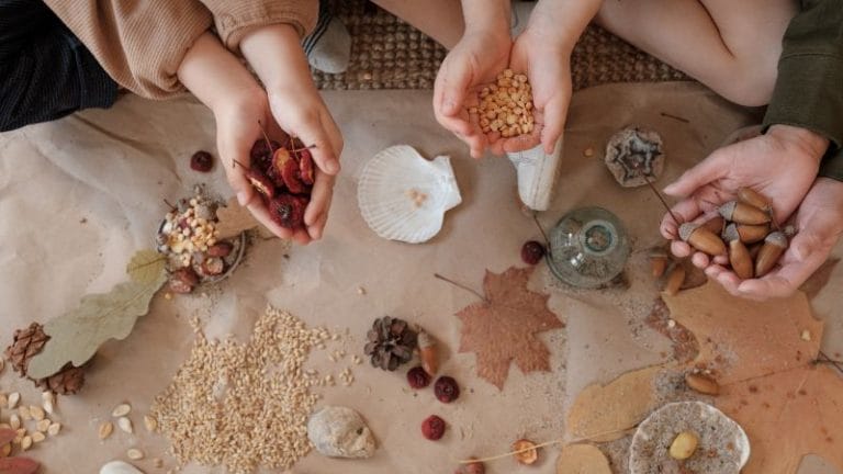 Three people sitting on the floor at a Nature-Based Home Daycare hold dried seeds, berries, and acorns in their hands above paper decorated with leaves, grains, and other natural objects.
