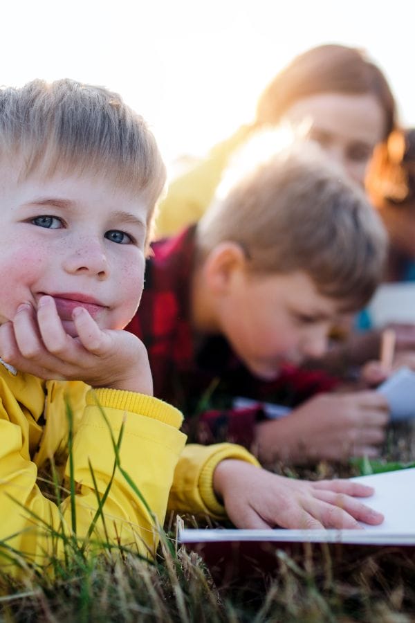 A boy in a yellow jacket lies on grass with an open notebook, looking at the camera, while other children write in the background at a Nature-Based Home Daycare.