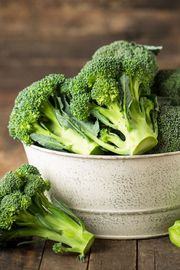 A white bowl filled with fresh broccoli florets sits on a wooden surface, with one floret resting outside the bowl—perfect inspiration for those looking to grow broccoli in Oklahoma.