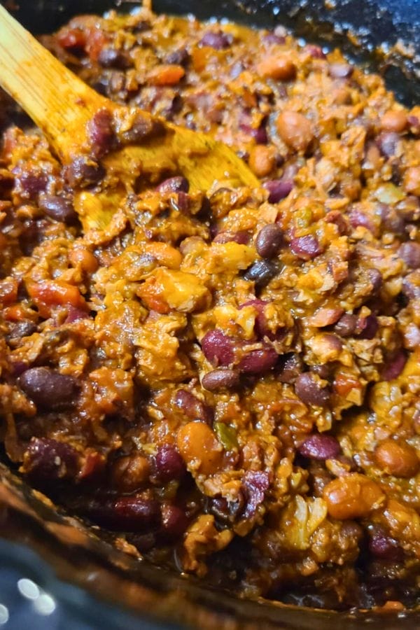 Close-up of a wooden spoon stirring a thick Leftover Brisket Chili mixture with beans, tomatoes, and ground meat in a pot.