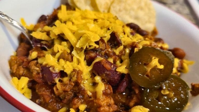 A bowl of Leftover Brisket Chili topped with shredded cheddar cheese, sliced jalapeños, and served with tortilla chips on the side.