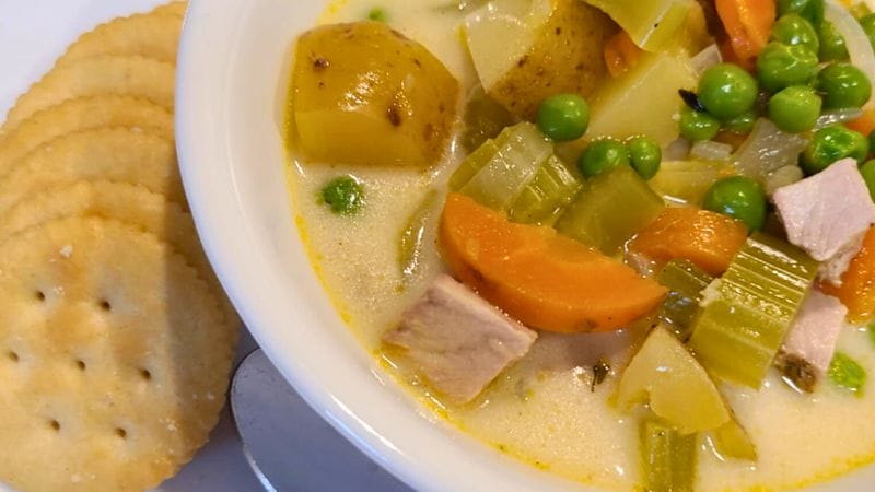A bowl of creamy soup with potatoes, carrots, celery, peas, and chunks of meat, served with round crackers on the side.