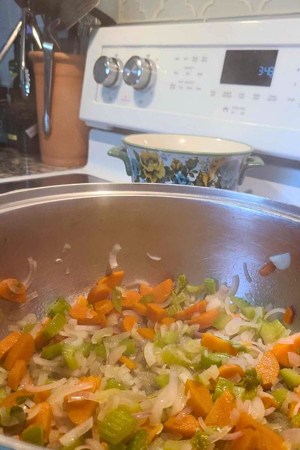 Chopped carrots, celery, and onions being sautéed in a pan on a stove, with a pot in the background—perfect veggies for starting a hearty Leftover Pork Chop Soup.