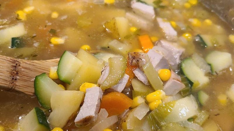 A close-up of Leftover Pork Chop Soup with diced zucchini, carrots, corn, and chunks of pork in broth, being stirred with a wooden spoon.