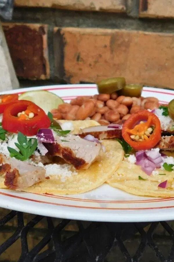 A plate of tacos topped with pork, red onions, cheese, parsley, and sliced peppers—perfect for Leftover Pork Chop Recipes—is served with pinto beans and jalapeño slices on a black metal table.