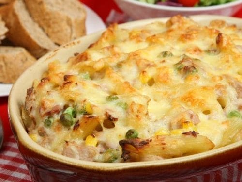 Baked pasta casserole with melted cheese, peas, and corn in a dish—one of our favorite freezer friendly tuna recipes—served alongside slices of brown bread and a partial view of salad in the background.