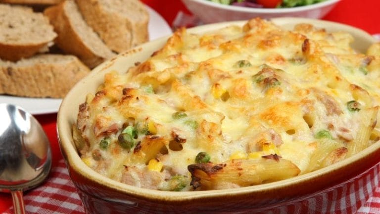 Baked pasta casserole with melted cheese, peas, and corn in a dish—one of our favorite freezer friendly tuna recipes—served alongside slices of brown bread and a partial view of salad in the background.