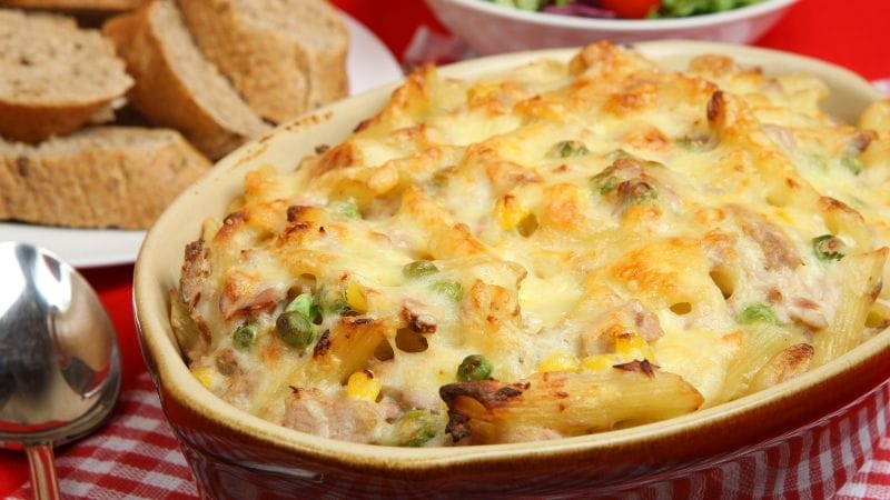 Baked pasta casserole with melted cheese, peas, and corn in a dish—one of our favorite freezer friendly tuna recipes—served alongside slices of brown bread and a partial view of salad in the background.