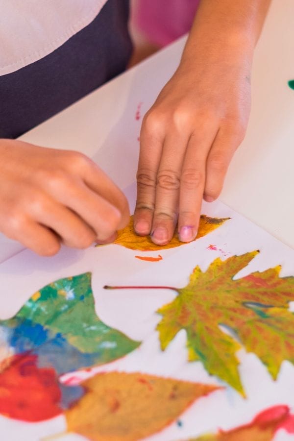 A person’s hands pressing a dried leaf onto paper, surrounded by colorful painted leaves, captures the creativity of crafts using leaves in this vibrant arts and crafts scene.
