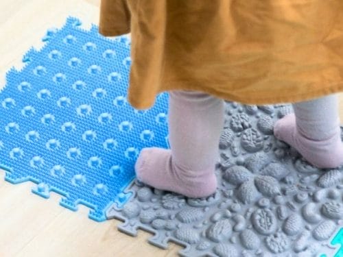 A child stands on textured interlocking foam mats, wearing light-colored tights and a brown outfit—perfect for inspiring DIY Sensory Play Ideas.