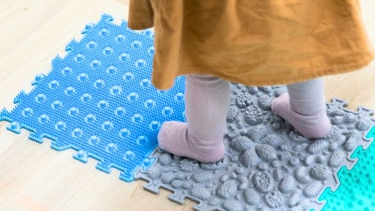 A child stands on textured interlocking foam mats, wearing light-colored tights and a brown outfit—perfect for inspiring DIY Sensory Play Ideas.