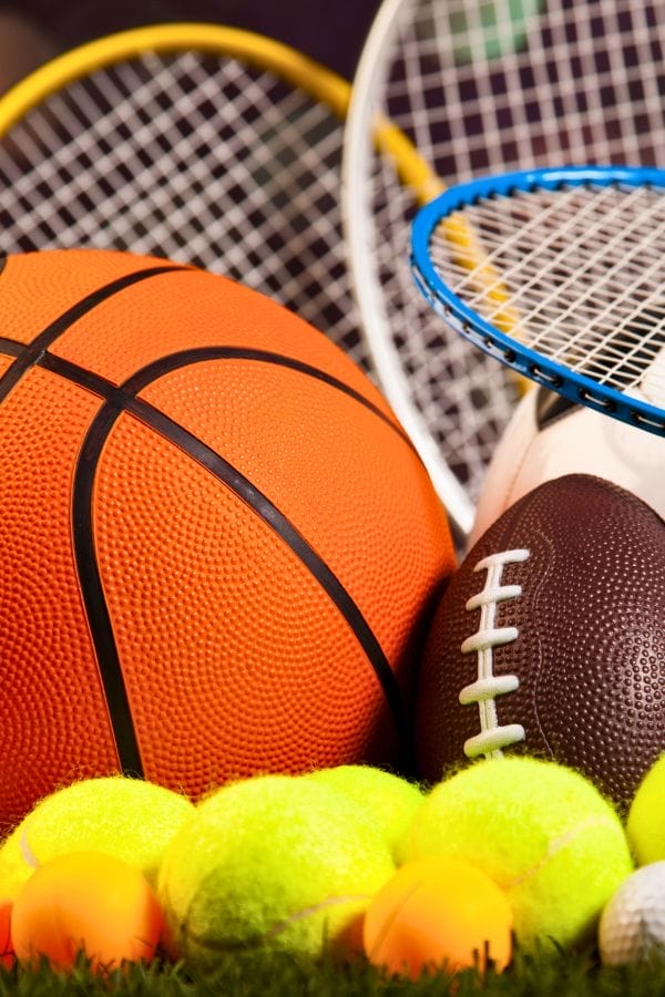 Assorted sports equipment, including a basketball, football, tennis balls, badminton racket, and tennis racket, arranged together on grass—perfect inspiration for your next Sports Shrinky Dink project.