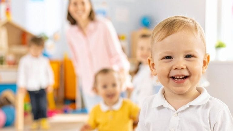 Smiling toddler in a white shirt stands in the foreground of a brightly lit room, with other children and an adult in the background—perfect for Childcare Advertising showcasing joyful, welcoming environments.