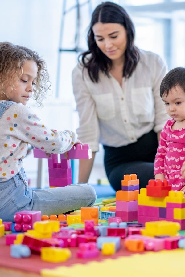 An adult and two children play with colorful plastic building blocks on a table in a bright room, capturing a joyful moment perfect for childcare advertising.