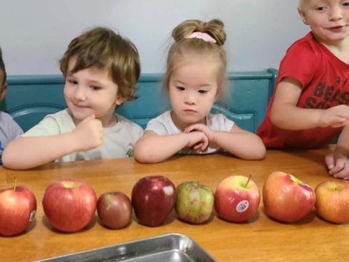 Four children sit at a table behind a row of assorted apples, ready to explore fun lesson plans on apples.