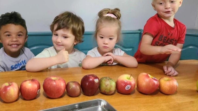 Four children sit at a table behind a row of assorted apples, ready to explore fun lesson plans on apples.
