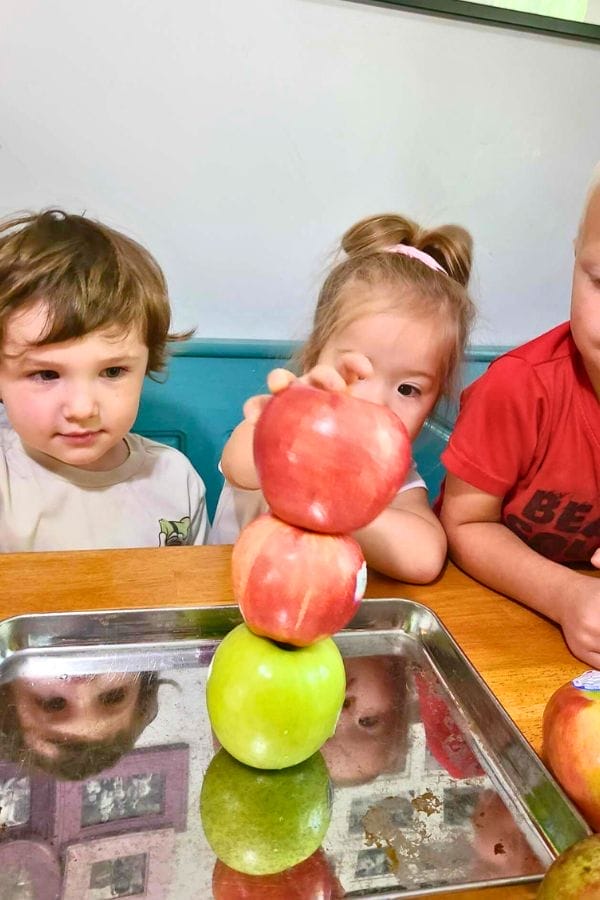 Three children sit at a table, watching as one of them stacks two red apples on top of a green apple on a metal tray—an engaging moment inspired by lesson plans on apples.