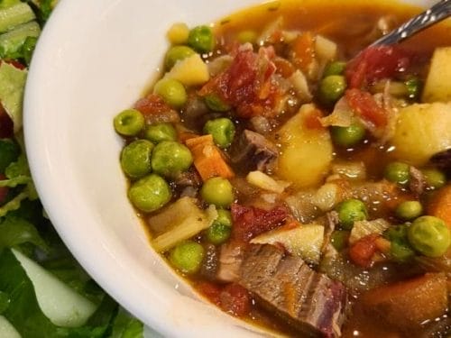 A bowl of vegetable beef stew with potatoes, carrots, peas, and tomatoes next to a side salad with lettuce and cherry tomatoes.