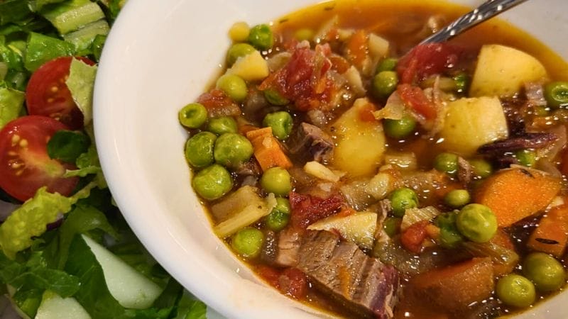 A bowl of vegetable beef stew with potatoes, carrots, peas, and tomatoes next to a side salad with lettuce and cherry tomatoes.