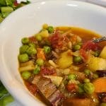 A bowl of vegetable and beef soup with peas, carrots, potatoes, and tomatoes. Fresh cherry tomatoes in a container are visible in the background.