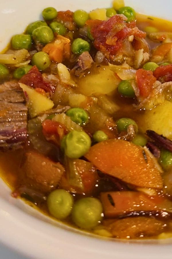 A close-up of a bowl of beef stew with peas, carrots, potatoes, tomatoes, and chunks of beef in a broth.