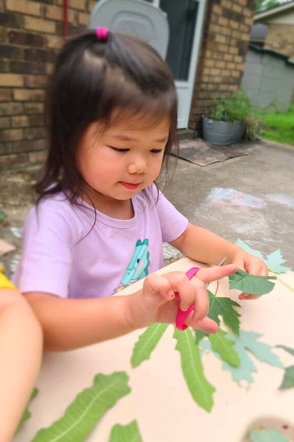 A young child in a purple shirt engages in educational activities with nature, using scissors to cut green leaves while sitting at a table outdoors.