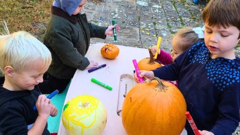 Four young children enjoy painting pumpkins with markers at an outdoor table. Various colored markers and pumpkins are scattered across the table.