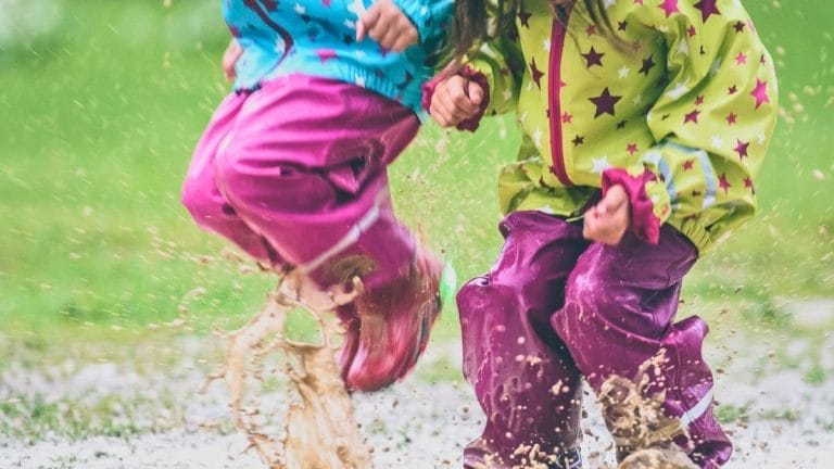 Two children wearing colorful rain gear enjoy weather activities in the garden, jumping into a muddy puddle and splashing water and mud around them on a rainy day.