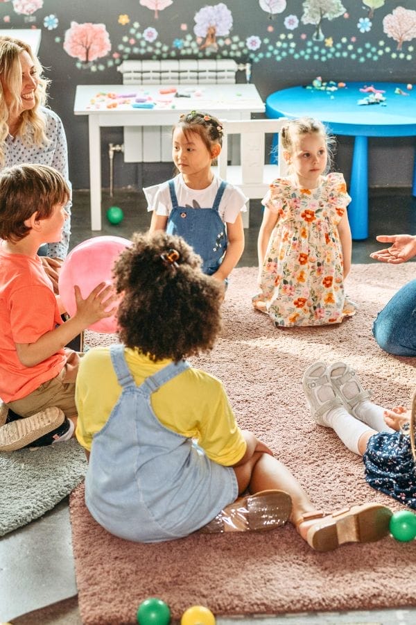 A group of young children sit in a circle on a rug in a classroom, with an adult nearby. Colorful decorations and toys fill the background, showcasing budget friendly daycare setups that create a cheerful and engaging space.