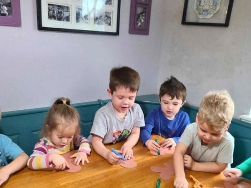 Six young children sit at a table, coloring paper shapes inspired by lesson plans on gingerbread with markers and crayons in a well-lit room decorated with framed pictures.