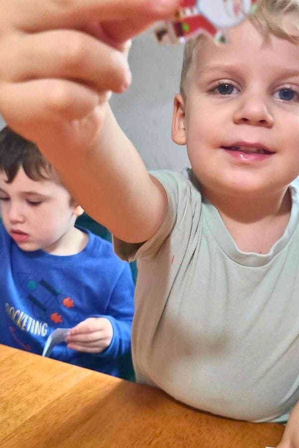 Two young children sit at a table; one is smiling and holding something up to the camera, while the other is focused on an activity—perfect inspiration for lesson plans on gingerbread.