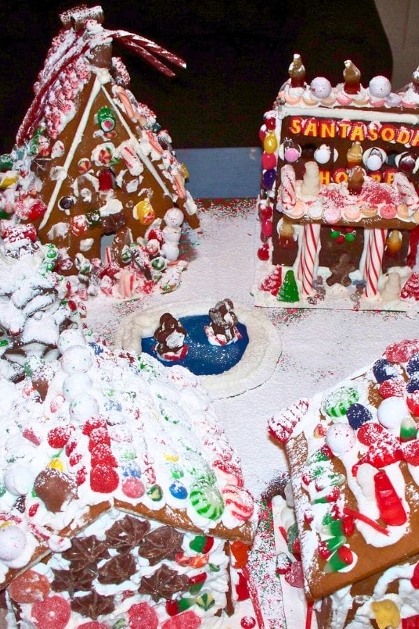 Several decorated gingerbread houses surround a small candy rink with two penguin figurines, all dusted with powdered sugar—perfect inspiration for creative lesson plans on gingerbread.