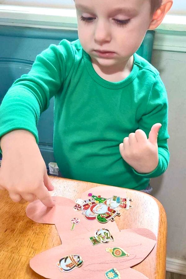 A young child in a green shirt places stickers on a pink paper cutout while sitting at a wooden table, inspired by lesson plans on gingerbread.