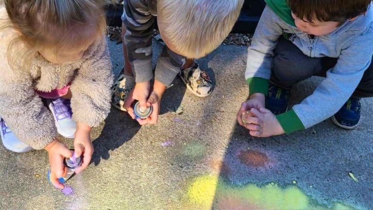 Three children crouch on a sidewalk, spraying colorful paint onto the concrete surface, inspired by lesson plans on Jean-Michel Basquiat.