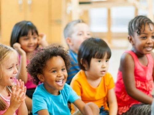 A group of young children sit together indoors, smiling and looking toward the front of the room, some clapping and appearing engaged during lesson plans on "Llama Llama Red Pajama.