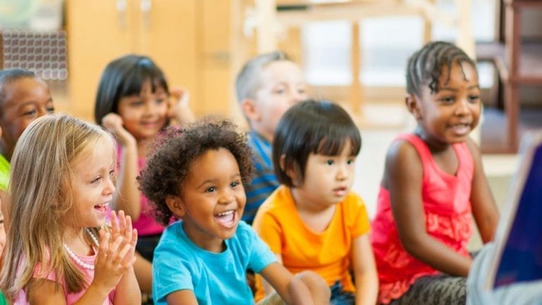 A group of young children sit together indoors, smiling and looking toward the front of the room, some clapping and appearing engaged during lesson plans on "Llama Llama Red Pajama.