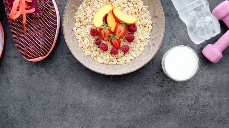 A bowl of oats with fruit, a glass of milk, pink dumbbells, a water bottle, and running shoes on a dark surface inspire healthy snacks and simple movement for your daily routine.