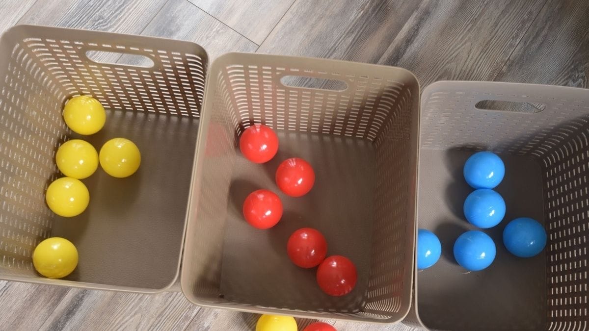 Three baskets on a wooden floor contain homemade toys: six yellow balls in the left basket, five red balls in the middle, and five blue balls in the right basket.