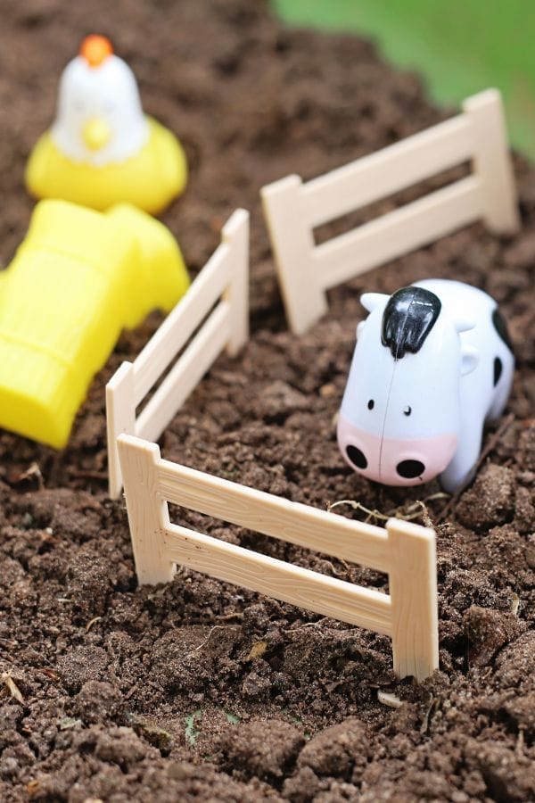 Homemade toys set the scene: a plastic toy cow stands in a small fenced area on soil, with a yellow toy chicken and barn in the background.