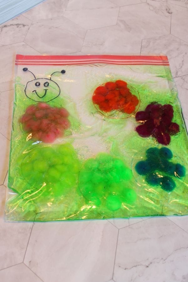 A clear plastic bag filled with colored pom-poms and gel, divided into sections, features a cartoon face and sits on a tiled surface—one of the many fun homemade toys for kids to enjoy.