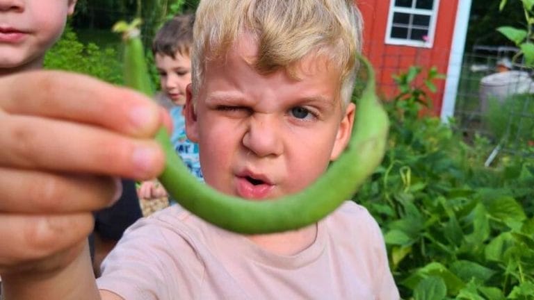 A young boy, engaged in Nature Based Learning, holds a curved green bean close to the camera, squinting one eye, with other children and garden plants visible in the background.