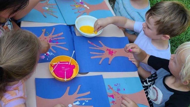 Five children sit around a table outdoors, enjoying Outdoor Art in Nature as they decorate handprint art with paint and cotton swabs on blue paper.
