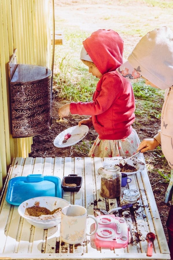 Two children wearing hooded jackets play with toy dishes and dirt at an outdoor play kitchen next to a yellow wall, enjoying hands-on Outdoor Science Activities.