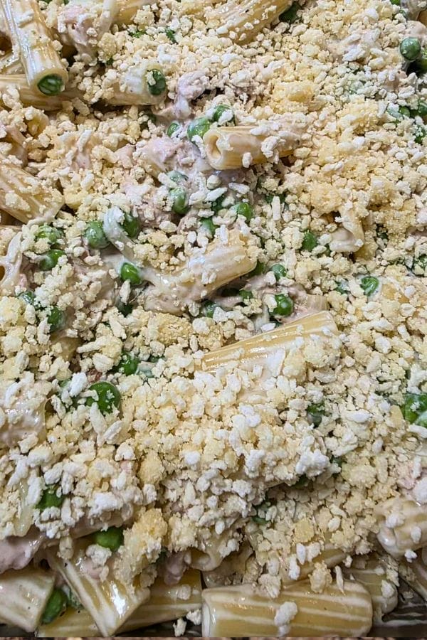 Close-up of a Tuna Alfredo Bake casserole dish with rigatoni pasta, green peas, and a crumbly topping, possibly made of crushed crackers or breadcrumbs.