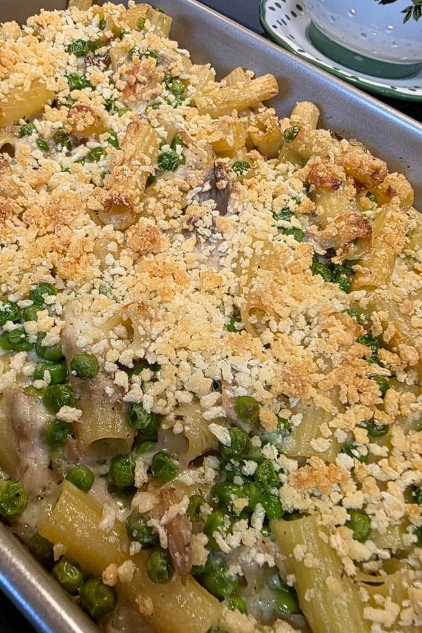A Tuna Alfredo Bake casserole topped with golden breadcrumbs, featuring rigatoni, green peas, mushrooms, and a creamy Alfredo sauce in a rectangular metal pan.