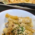 A bowl of baked rigatoni pasta with peas and breadcrumbs, reminiscent of a comforting Tuna Alfredo Bake, sits in front of a baking dish filled with the same hearty dish.
