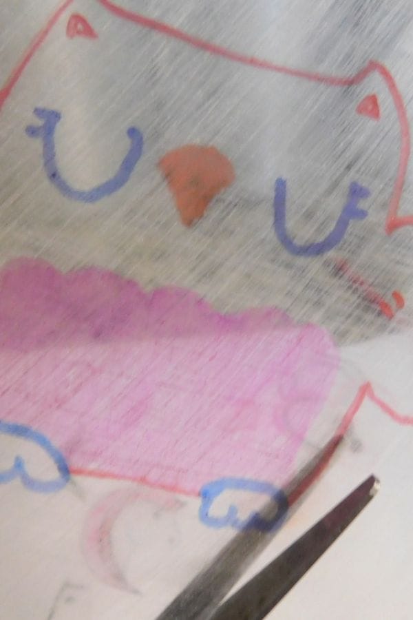 A close-up photo of a cartoon owl drawing, perfect for Winter Shrinky Dink Patterns, with closed eyes, an orange beak, pink belly, and blue feet on a shiny, scratched surface. Scissors appear at the bottom right.