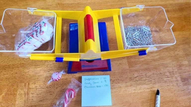 A yellow balance scale on a table weighs a cookie on one side and metal clips on the other, with candy, a pen, and a note listing weights—perfect props for fun Christmas Math Activities.